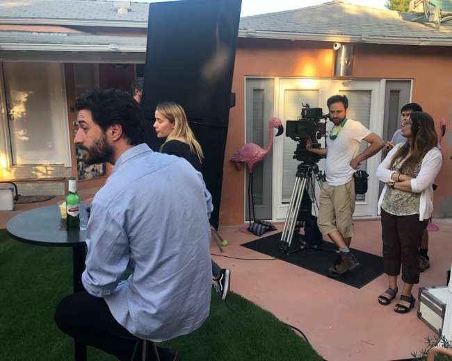 A film crew sets up a scene in an outdoor area with a man seated at a table and additional crew members observing and operating equipment.