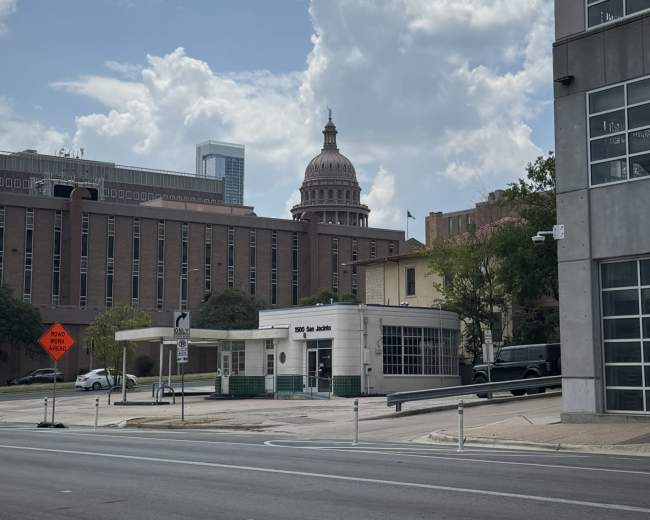 The Texas State Capitol dome is visible in the background, surrounded by several buildings and a clear blue sky with clouds.