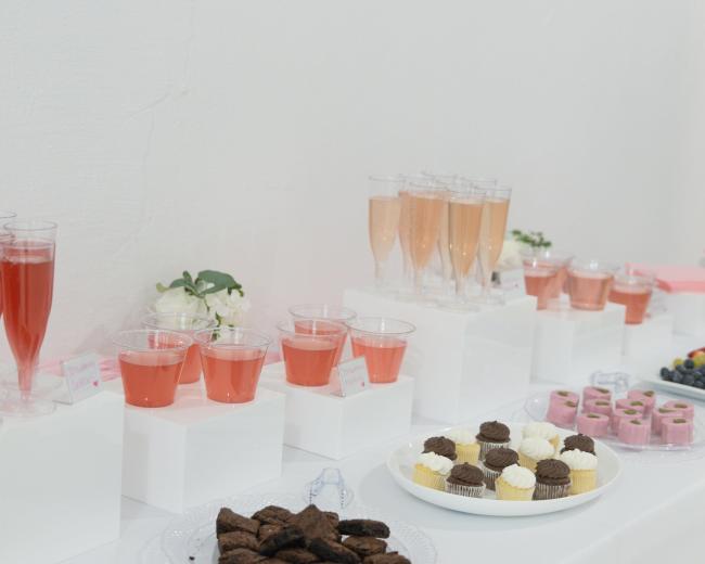 A table displaying various drinks in clear glasses, including pink beverages, alongside an assortment of desserts such as cupcakes, cookies, and gelatin treats.