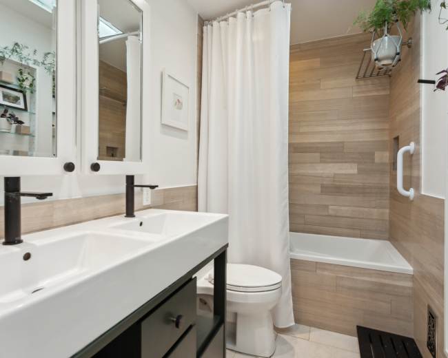 The image shows a modern bathroom featuring a double sink vanity, a white bathtub with a shower curtain, and wooden wall accents.