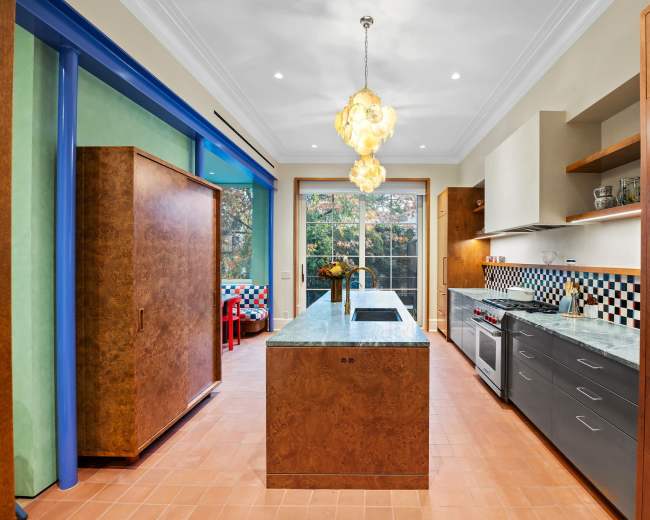 The modern kitchen features a large island with a stone countertop, wooden cabinetry, and colorful decorative elements, with large windows allowing natural light to fill the space.