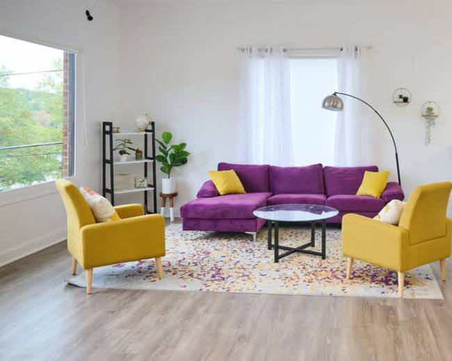 A modern living room features a purple sectional sofa, two yellow accent chairs, a circular glass coffee table, and a decorative area rug with a floral design.