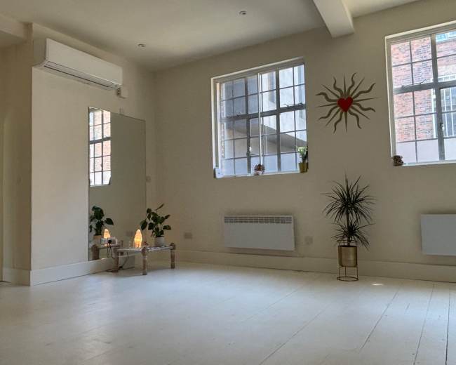 The image shows a spacious, warmly lit room with large windows, a mirror, plants, and decorative elements including a heart inside a sun design on the wall.
