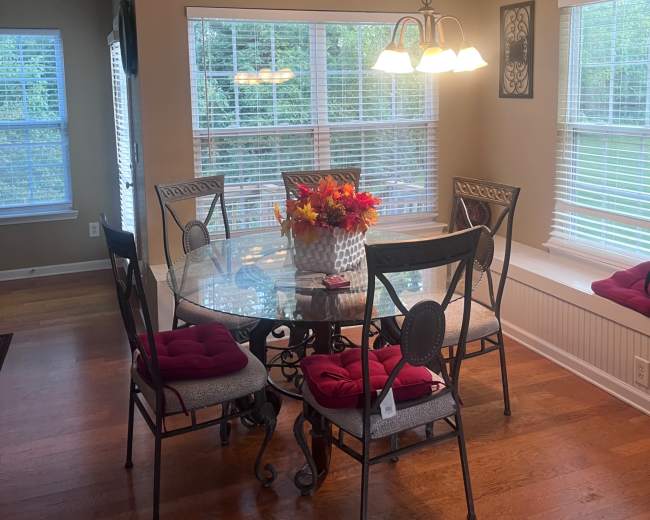 A glass dining table surrounded by six chairs with red cushions is illuminated by a pendant light in a room with multiple windows.