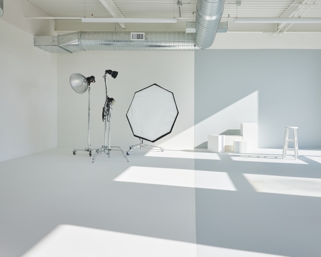 The image shows a bright photography studio with a large softbox, a stand for a light, and a small white stool, all positioned on a light-colored floor.