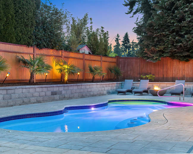 A well-lit outdoor pool area with lounge chairs and palm trees surrounded by a wooden fence.