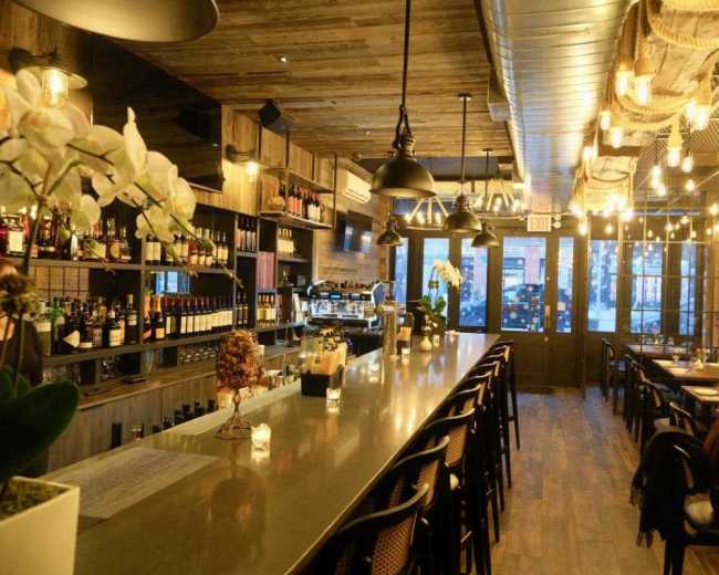 The image shows a modern restaurant interior with a long bar, shelves filled with bottles, and a few patrons seated at tables.