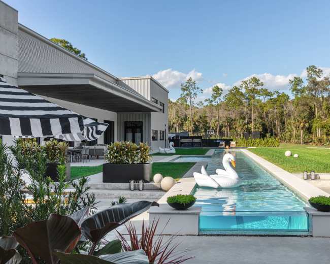 A modern backyard features a rectangular swimming pool with a swan float, surrounded by lush green grass, lounge chairs, and patio furniture under a striped awning.