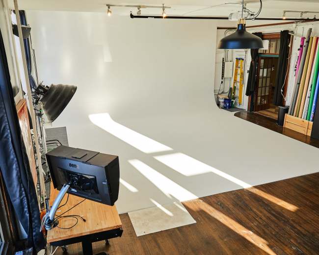 A photography studio with a large white backdrop, wooden flooring, and various lighting and equipment arranged around the space.