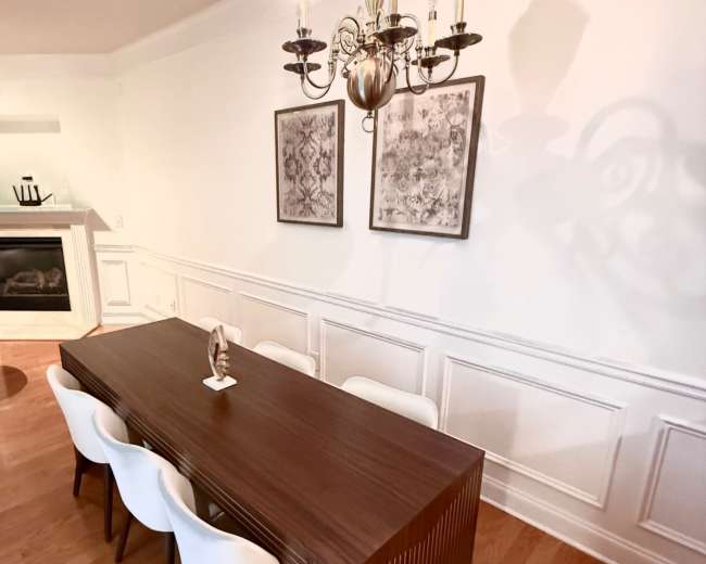 A long, wooden dining table with white chairs is positioned under a chandelier in a well-lit room featuring decorative wall panels and two framed artworks.