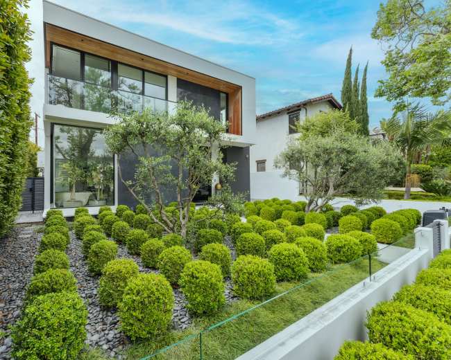 A modern house with large windows is surrounded by neatly manicured green hedges and stone pathways.