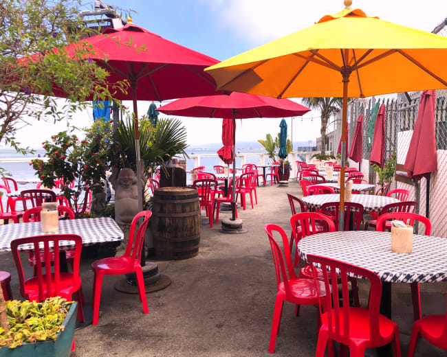 A patio area features red plastic chairs and tables, shaded by brightly colored umbrellas, alongside a water view.