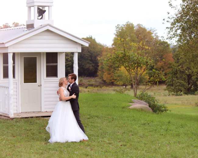 A couple stands embracing in front of a small white building on a grassy area with trees in the background.