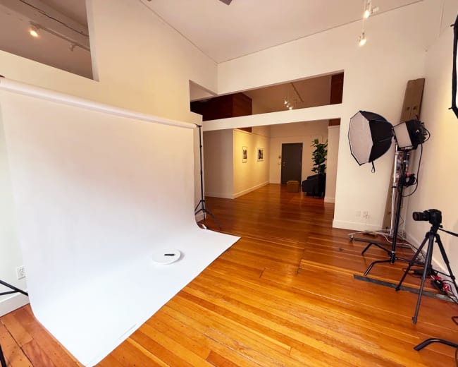 The image shows a photography studio with a white backdrop, wooden floors, and several lighting equipment set up.