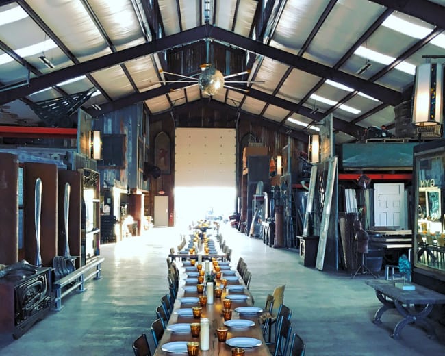The scene shows a long wooden dining table set with plates and amber glasses inside a spacious, industrial-style warehouse.
