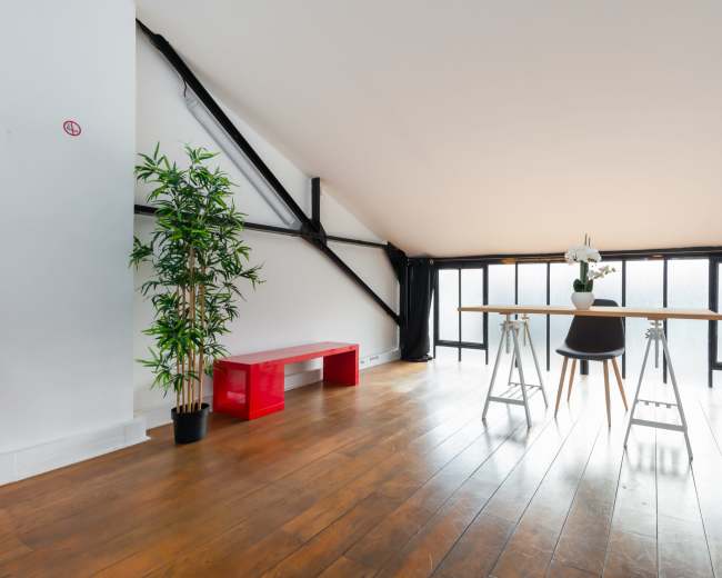 The image shows a bright, minimalist loft space with a wooden floor, a simple dining table, a plant, and large windows.