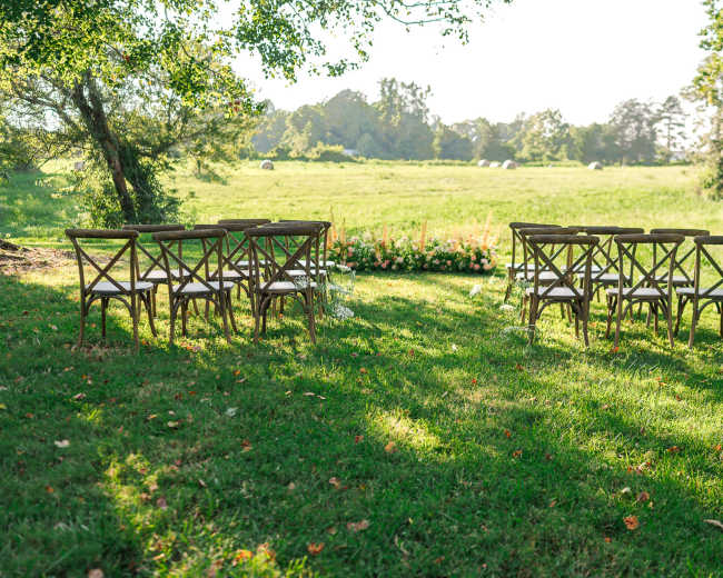 A row of wooden chairs is arranged on a grassy area facing a flower-lined aisle in a natural outdoor setting.