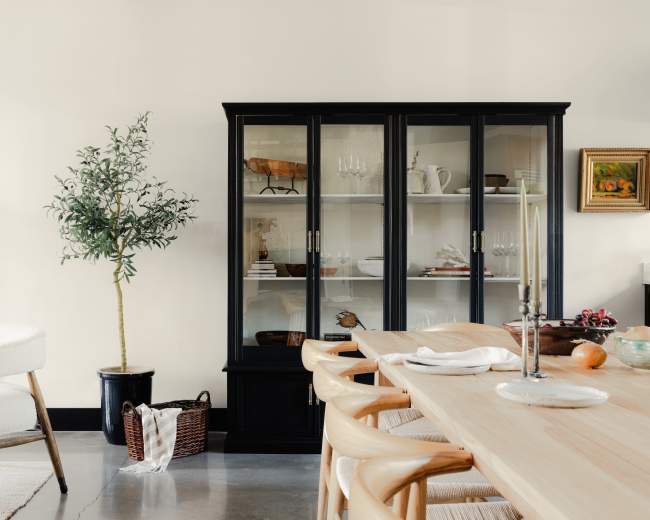 The image shows a modern dining area featuring a wooden table, a black glass-front cabinet displaying tableware, a potted plant, and minimalist decor on the walls.