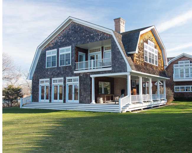 The image shows a large shingle-style house with multiple windows, a wraparound porch, and landscaped green lawns.