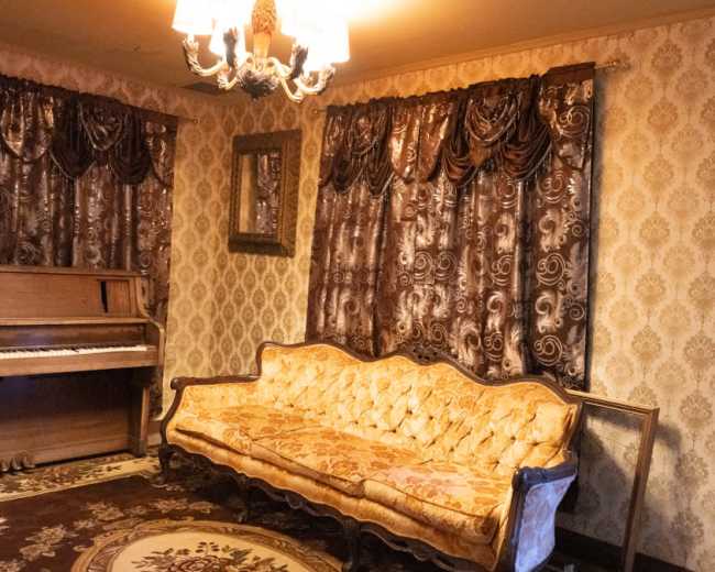 The room features a vintage piano, a decorative sofa with ornate upholstery, and patterned curtains against a floral wallpaper backdrop.