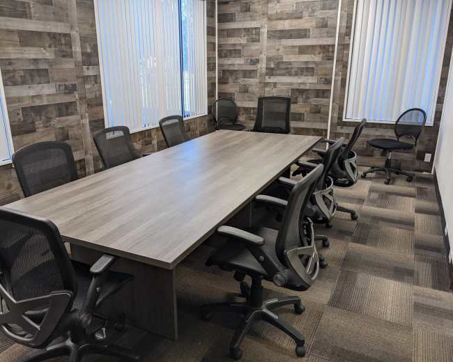 A conference room features a large rectangular table surrounded by black rolling chairs, with wooden walls and vertical blinds on the windows.