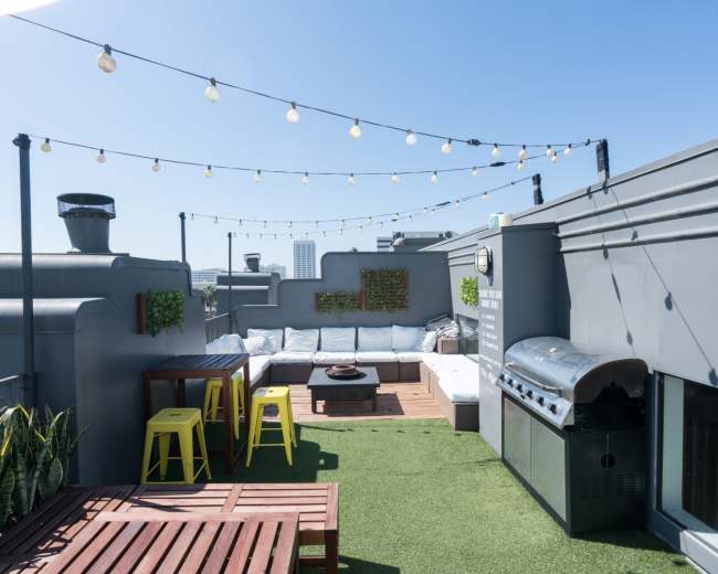The image shows a rooftop terrace with string lights, a grill, a seating area, and wooden furniture.