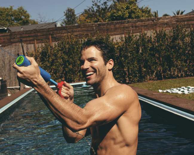 A shirtless man stands in a swimming pool, smiling while playfully aiming a water toy.
