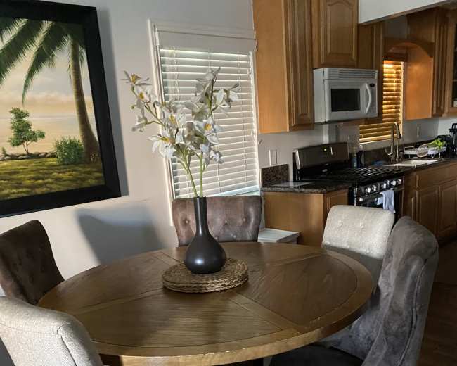 A round wooden dining table with four chairs sits in a well-lit kitchen area, accompanied by a vase of flowers on the table and a large painting of a landscape on the wall.
