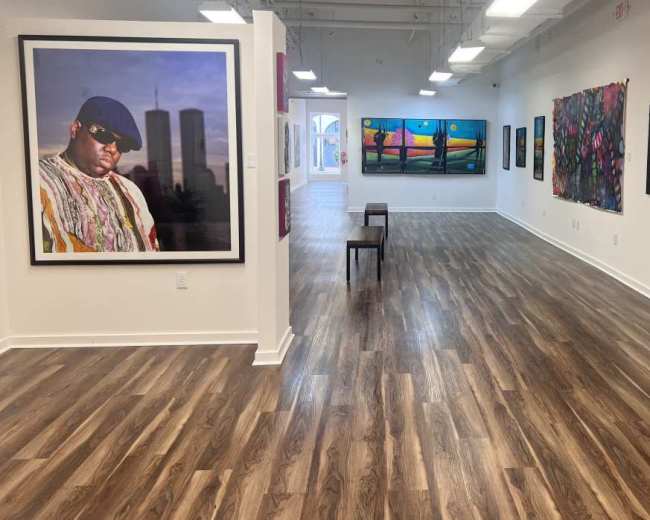 The image shows an art gallery featuring various paintings on the walls and a single chair positioned in the center of the room.