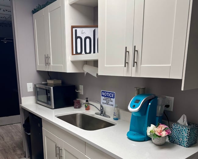 The image shows a modern kitchen with white cabinets, a stainless steel sink, a microwave, and a teal-colored coffee maker on a countertop.