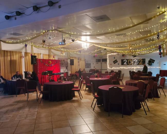 A banquet hall decorated with string lights and a stage set up for a performance, featuring round tables and chairs arranged for an event.