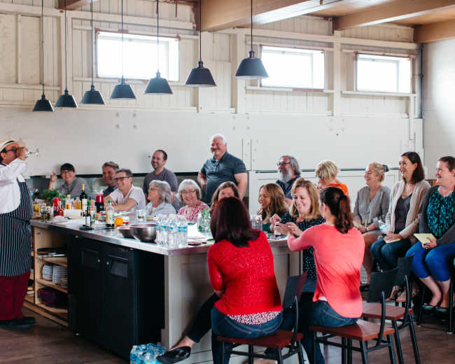 A chef demonstrates cooking techniques to a diverse audience seated at a kitchen island during a workshop.