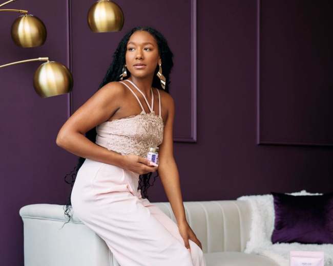 A woman in a light pink dress sits on a white sofa in front of a purple wall, holding a small jar while looking towards the camera.