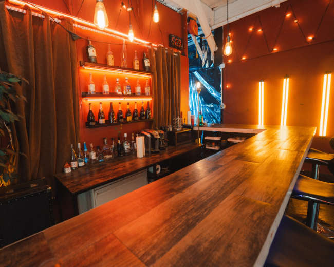 The image shows a well-lit bar area with a wooden counter, shelves displaying various bottles, and warm lighting accents.