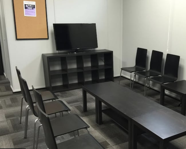 The image shows a small meeting room with a television, two tables, and several black chairs arranged around them.
