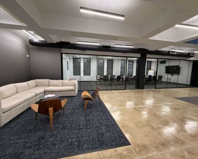 The image shows a modern office lounge area with a sofa, a coffee table, and a view of a glass-walled conference room.