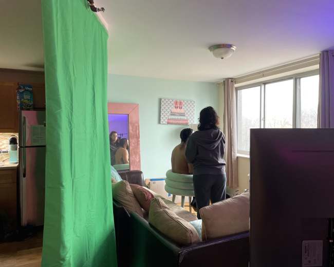 A person is seated in a chair while another person stands behind them, with a green screen and a television visible in a living room setting.