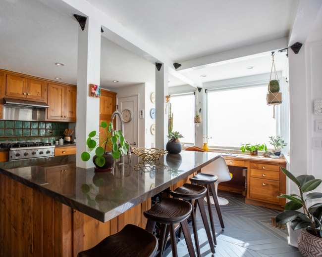 The image shows a kitchen with wooden cabinets, a large island with dark countertops, and a small workspace by a window, featuring plants and natural light.