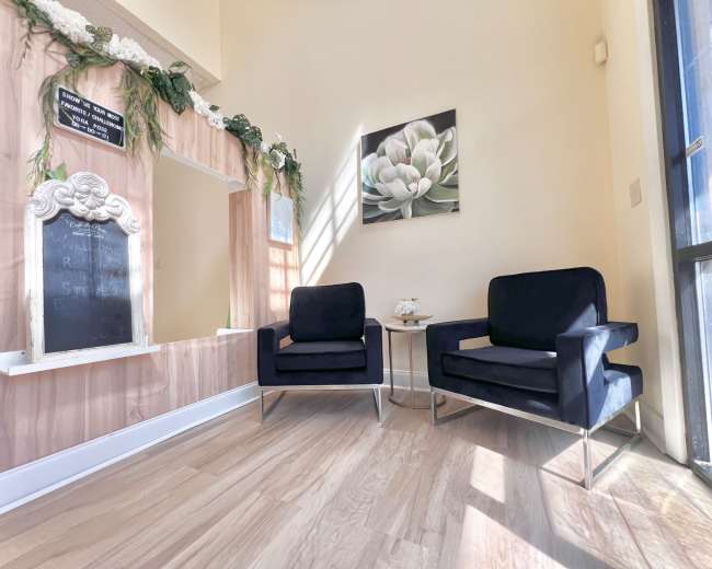 The image shows a well-lit interior space featuring two black upholstered chairs against a wall with a decorative mirror and potted plants.