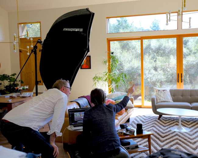 Two people are reviewing images on a laptop in a well-lit living room with large windows overlooking greenery.