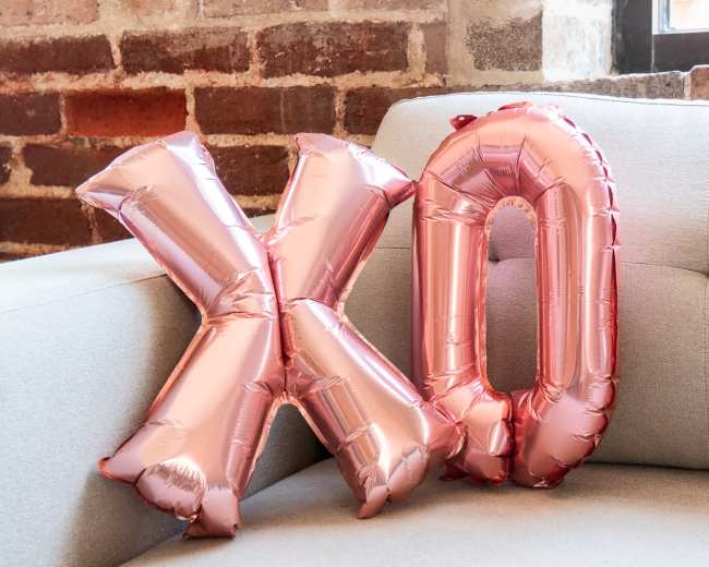 Two rose gold foil balloons shaped like the letters "X" and "O" rest against a light gray couch in front of a brick wall.