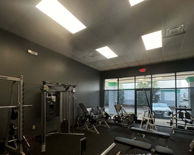 The image shows a fitness room with various exercise equipment, including weight benches, a cable machine, and stationary bikes, all arranged along the perimeter near large windows.