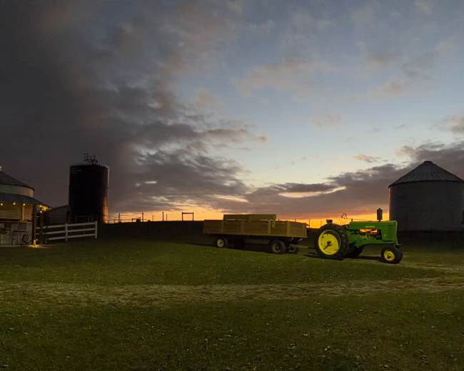 A green tractor and a trailer are positioned on a farm at dusk, with a barn and silos in the background under a cloudy sky.