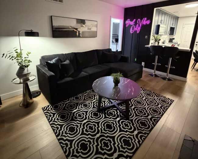 The living room features a black sofa and a round glass coffee table, with a patterned rug underneath and a neon sign on the wall.
