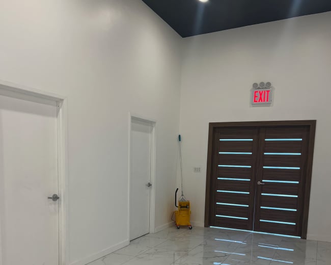 The image shows a bright, minimalist entryway with white walls, two doors on the left, and a brown double door with horizontal slats on the right, accompanied by a yellow cleaning bucket in the corner.
