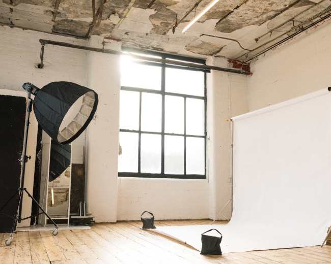 A photography studio features a large light source, a white backdrop, and wood flooring with exposed brick walls.