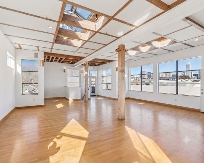 The image shows a spacious, well-lit room with wooden flooring, large windows, and a ceiling featuring exposed beams and skylights.