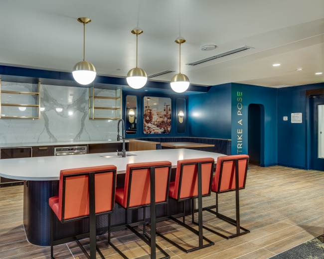 The image shows a modern kitchen area featuring a large white island with red bar stools, illuminated by pendant lights, against a blue wall with decorative shelves and a motivational phrase.