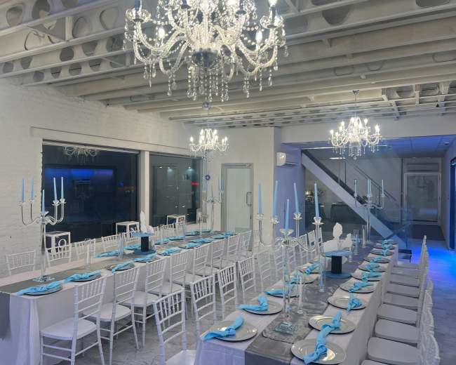 A long dining table is set with blue napkins and white chairs under chandeliers in a well-lit room.
