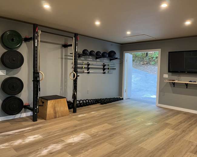 The image shows a home gym with weight plates, a squat rack, kettlebells, exercise balls, and a television mounted on the wall.